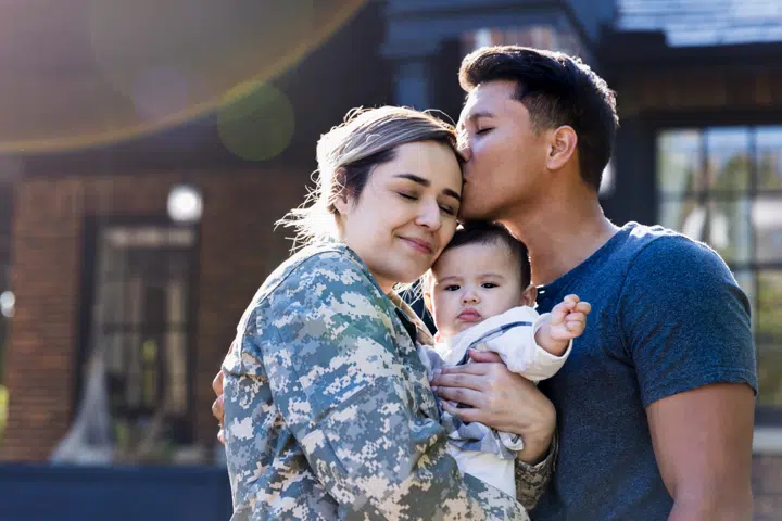 Military service member in uniform embraced by their partner and holding a baby, symbolizing support for veterans and military families.