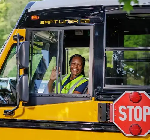Smiling bus driver waving from the driver’s seat of a yellow school bus, representing rewarding driver opportunities with comprehensive training and flexible shifts