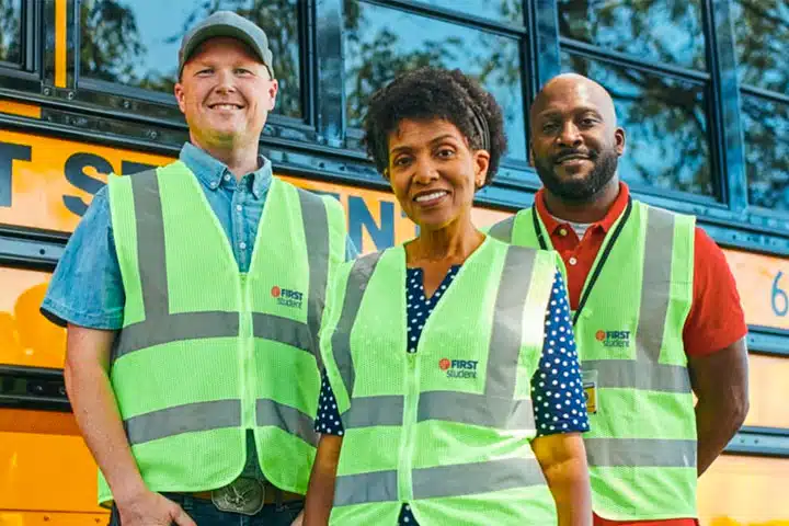 Three First Student employees wearing reflective safety vests standing in front of a yellow school bus, representing the company’s commitment to diversity and inclusion.