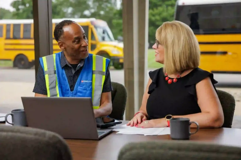 Two First Student employees having a discussion at a table with a laptop and papers, symbolizing leadership training and professional development opportunities.