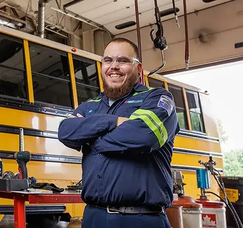 Smiling mechanic wearing safety glasses and a uniform standing in a bus maintenance garage, representing advanced training and technology opportunities for technicians.