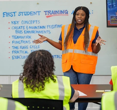 Instructor wearing an orange safety vest leading a training session for bus drivers in high-visibility vests, representing First Student’s commitment to smooth and safe operations.