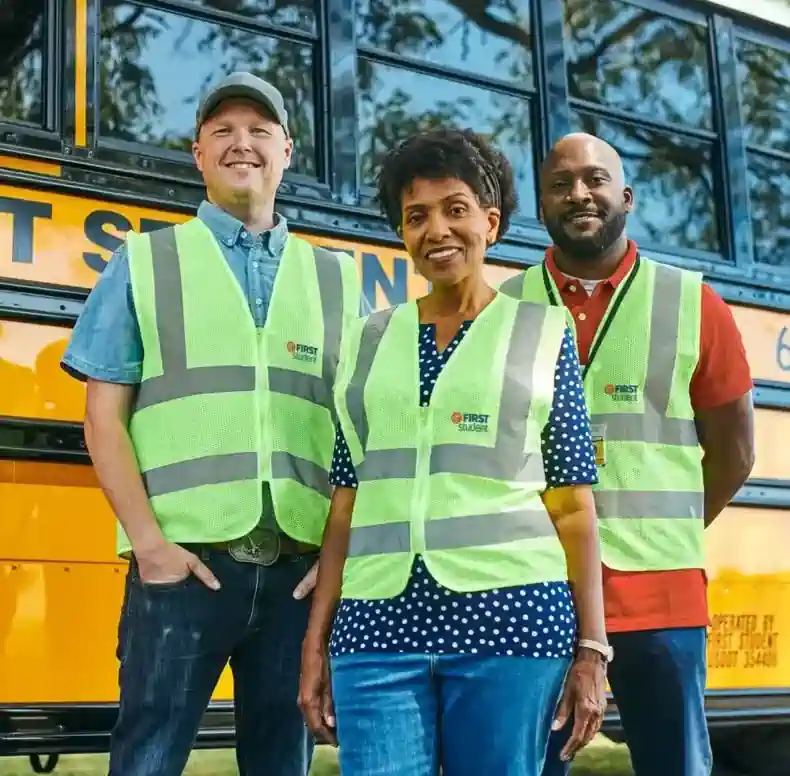 Three First Student employees wearing reflective safety vests standing in front of a yellow school bus, representing rewarding career opportunities for school bus drivers.